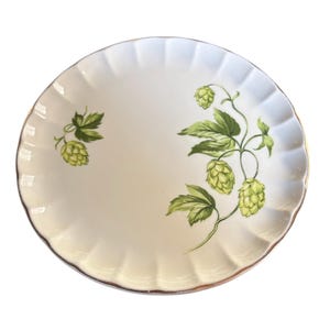 May include: A white ceramic plate with a scalloped edge and a gold rim. The plate is decorated with green hop plant illustrations. The hops and leaves are painted in detail, adding a touch of nature to the tableware.