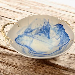 Op de afbeelding: Een ronde, cr&egrave;mekleurige serveerschaal met abstracte blauwe wervelingen. De kom heeft twee gevlochten touwhandvatten. Het interieurontwerp heeft een marmereffect met blauwtinten. De kom staat op een houten oppervlak.