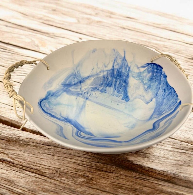 Op de afbeelding: Een ronde, cr&egrave;mekleurige serveerschaal met abstracte blauwe wervelingen. De kom heeft twee gevlochten touwhandvatten. Het interieurontwerp heeft een marmereffect met blauwtinten. De kom staat op een houten oppervlak.