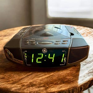 Puede incluir: Un radio reloj estéreo CD de doble alarma GE negro que muestra las 12:41 AM en números digitales verdes. El radio reloj tiene botones para snooze, sleep y presintonías de radio. Está sobre una superficie de madera.