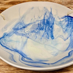 Op de afbeelding: Een witte keramische serveerschaal met een blauw gemarmerd ontwerp. De schaal heeft een afgeronde vorm met een licht verhoogde rand en twee kleine gaatjes om op te hangen. Het blauwe patroon wervelt over het oppervlak.