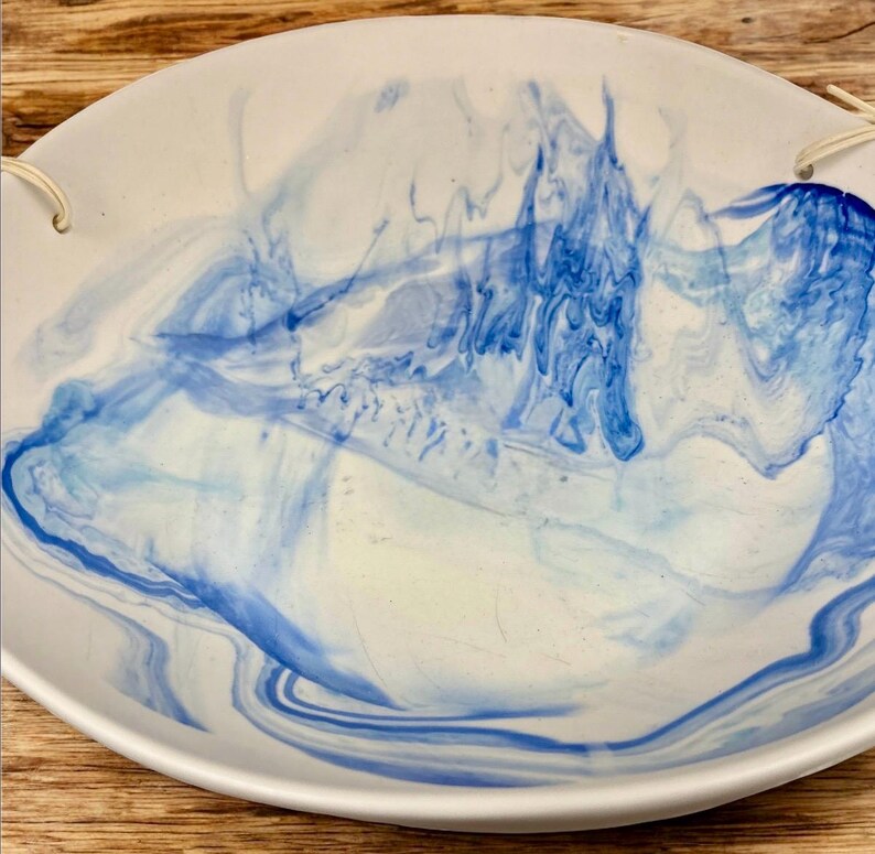 Op de afbeelding: Een witte keramische serveerschaal met een blauw gemarmerd ontwerp. De schaal heeft een afgeronde vorm met een licht verhoogde rand en twee kleine gaatjes om op te hangen. Het blauwe patroon wervelt over het oppervlak.