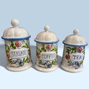 May include: A trio of ceramic kitchen storage jars with lids. Each white jar has a blue rim and is decorated with floral and ribbon motifs. The labels read "SUGAR", "COFFEE", and "TEA".