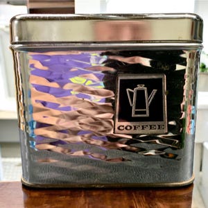 May include: A vintage silver-toned coffee tin with a textured, hammered finish. The rectangular container has a hinged lid and a black label with a coffee pot graphic and the word "COFFEE".