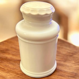 Puede incluir: Un tarro de cerámica blanco con tapa. El tarro tiene un cuerpo cilíndrico con una base redondeada y un cuello ligeramente más estrecho. La tapa tiene un diseño festoneado. El tarro está sobre una superficie de madera.