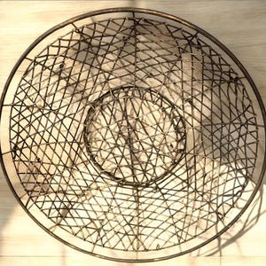 以下が含まれることがあります： 織り込まれたワイヤーデザインの、丸いアンティークスタイルの金属製バスケット。バスケットは、中央が盛り上がった丈夫なリムが付いた円形です。金属は素朴で古びた外観をしており、ヴィンテージまたはヴィンテージ風であることを示唆しています。