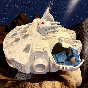 Puede incluir: Un detallado modelo de juguete blanquecino de la nave espacial Halcón Milenario, con intrincados paneles, torretas y un acento rojo. El interior de la nave es visible con detalles azules. El modelo está ambientado sobre un fondo de cielo estrellado y terreno rocoso.