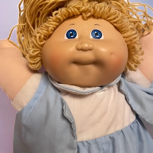 Può includere: Una bambola vintage con un viso rotondo, occhi blu e capelli biondi di filo. La bambola indossa un vestito azzurro con un colletto bianco. Le guance della bambola sono rosee e ha un leggero sorriso.