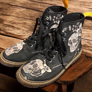 Puede incluir: Botas de lona negras con un diseño de calaveras y flores. Las botas tienen cierre de cordones, punta redonda y suela marrón. El diseño incluye una calavera, rosas y otras flores en tonos grises y blancos.