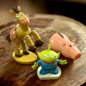 May include: Three character figurines from the animated film Toy Story. A light green alien with three eyes, a pink piggy bank, and a yellow donkey figurine with a brown saddle are displayed on a wooden surface.