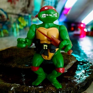 Puede incluir: Figura de acción verde de Rafael de las Tortugas Ninja. La figura lleva una bandana roja, armadura amarilla en el pecho y la cintura, y un cinturón negro. Sostiene un arma marrón y está sobre una superficie de piedra.