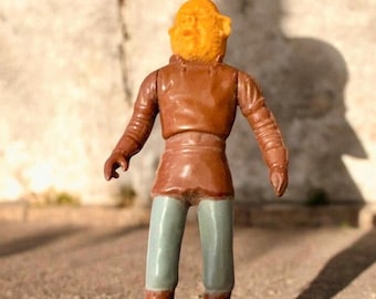 May include: A vintage action figure of a character with a yellow head and brown outfit. The figure has a brown jacket, light green pants, and brown boots. The figure is standing in front of a textured wall.