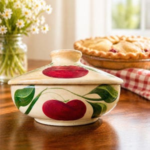 Puede incluir: Un cuenco de cerámica color crema con tapa, adornado con cerezas rojas y hojas verdes pintadas a mano. El cuenco está sobre una superficie de madera, con una tarta y flores en el fondo. El cuenco se utiliza probablemente para servir o guardar alimentos.