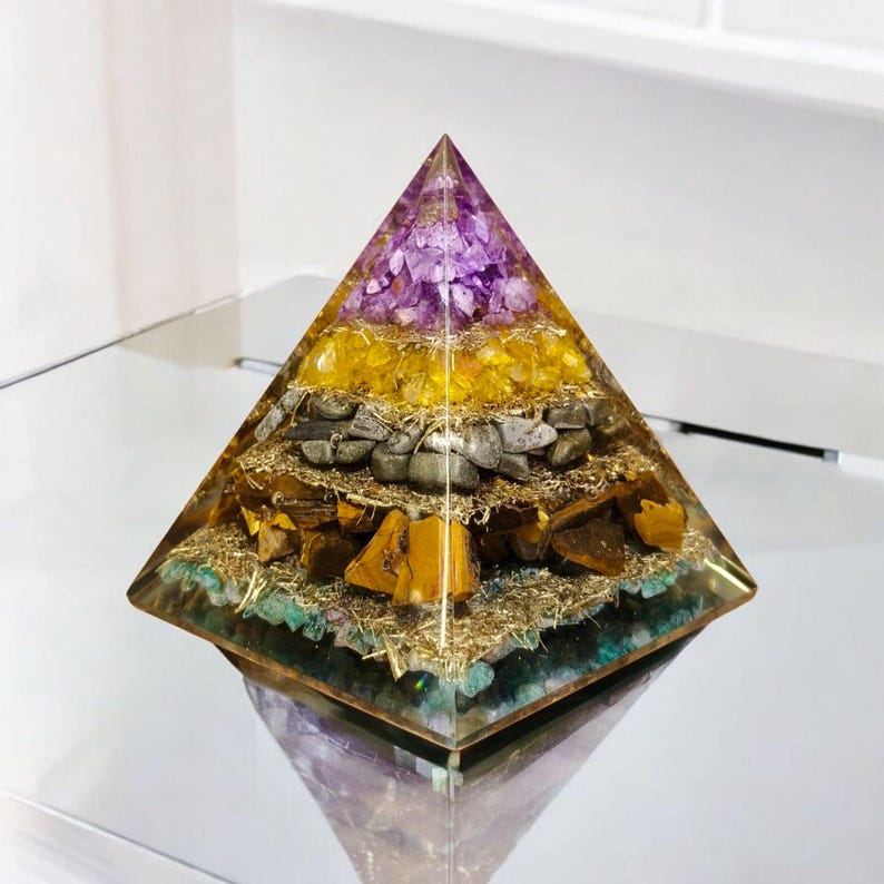 May include: A layered pyramid-shaped decorative object. The pyramid is made of clear resin and contains layers of amethyst, yellow stones, gray stones, and tiger's eye. The base has a turquoise hue, and the pyramid is resting on a reflective surface.