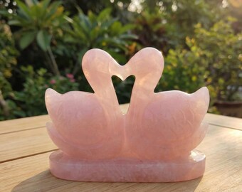 May include: A pink rose quartz sculpture of two swans facing each other, creating a heart shape. The swans have detailed feather carvings and rest on a rectangular base. The sculpture is displayed on a wooden surface.