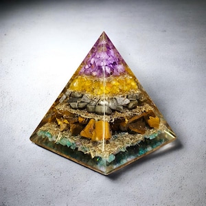 May include: A layered pyramid sculpture with a clear resin exterior. The pyramid features layers of purple, yellow, gray, and brown stones, with gold-colored accents. The base has a turquoise and green hue.