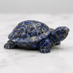 Può includere: Una statuetta di tartaruga in lapislazzuli scolpito. La scultura blu scuro presenta macchie dorate e poggia su una superficie bianca. Il guscio e il corpo della tartaruga sono finemente dettagliati, mostrando i motivi della pietra naturale.