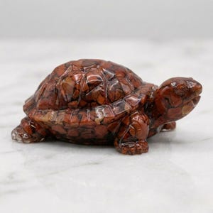 Puede incluir: Figurita de tortuga de piedra preciosa marrón rojiza detallada. La tortuga tiene un caparazón texturizado y está tallada con intrincados detalles. La figura es un artículo decorativo, probablemente para exhibición.
