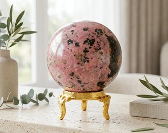 May include: A polished rhodonite sphere featuring a mottled pattern of soft pink and deep black mineral inclusions. The stone sphere rests securely upon a decorative, three-legged gold-toned metal stand with ornate, curved feet and a textured finish.