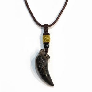 Può includere: Collana con cordino marrone scuro con un ciondolo nero scolpito a forma di dente di lupo. Il ciondolo è sospeso a un cordino con un nodo decorativo, una perla color oro e una perla nera.
