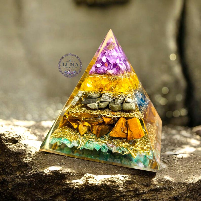 May include: A layered pyramid-shaped object with a clear resin exterior. The layers contain various crystals in shades of purple, yellow, orange, and green, along with gold-colored accents. The pyramid is displayed on a stone surface.