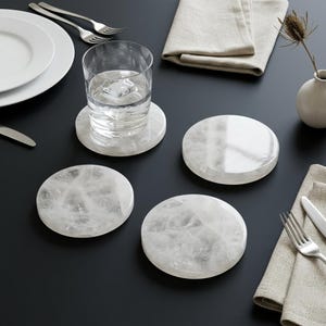 May include: A set of four circular coasters crafted from white stone with a polished, translucent finish. Each coaster features a unique, natural veined pattern throughout the surface, providing a smooth, reflective texture for drink placement.