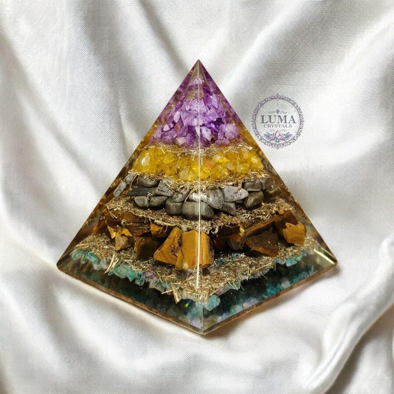 May include: A layered pyramid sculpture with a clear resin exterior. The pyramid contains layers of purple, yellow, gray, and brown stones. The base has a teal and gold color. The pyramid is on a white satin surface.