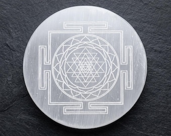 Peut inclure: Une dalle de pierre sélénite blanche circulaire présentant un motif géométrique Sri Yantra finement gravé. La surface de la pierre affiche une texture fibreuse naturelle avec un éclat subtil, contrastant avec les lignes fines et précises.