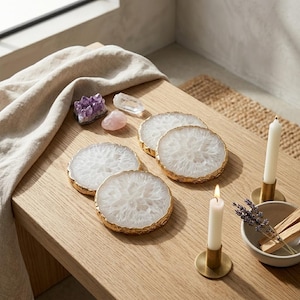 May include: A set of white agate coasters with gold rims, displayed on a light wooden table. The coasters have a circular shape and a natural, crystal-like design. Other items include crystals, candles, and a small bowl with dried herbs.
