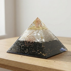 May include: An orgone energy pyramid with a layered design. The pyramid is made of black, white, and gold materials set in clear resin. A copper coil and spiral are visible. It rests on a wooden surface.