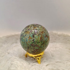 Peut inclure: Une sphère de pierre polie avec un motif moucheté vert, marron et violet. La sphère repose sur un petit support en métal doré à trois pieds ornés. L'arrière-plan est une surface douce, blanche et duveteuse.