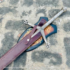 May include: A silver sword with an ornate handle and crossguard is sheathed in a burgundy leather scabbard. The scabbard rests on a decorative wooden plaque with gold detailing. The sword's blade is not visible. The plaque is dark blue and brown.