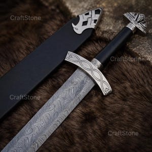 May include: A sword with a patterned Damascus steel blade, a black handle, and a silver-coloured pommel and crossguard featuring an infinity knot design. The sword includes a black scabbard with a silver-coloured metal tip featuring a lattice pattern.