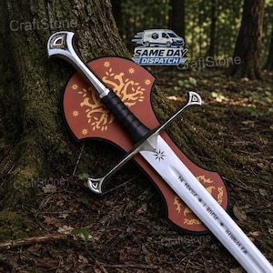 May include: A silver-toned metal sword with a black leather-wrapped grip and a crossguard, featuring etched runic symbols on the blade. The sword rests against a red wooden wall plaque decorated with a gold-coloured tree and star pattern.