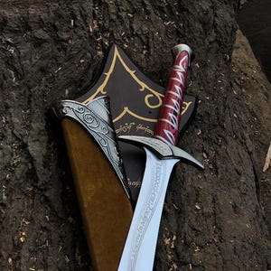 May include: A decorative sword with a red and silver handle, in a brown sheath. The silver blade has etched text. The sheath is set against a dark brown wooden plaque with gold accents and text.