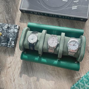 May include: A green leather watch roll holding three luxury watches with silver and black watch faces. A book titled "A Man & His Watch" is visible in the background. A marble case and a green wallet are also in the image.