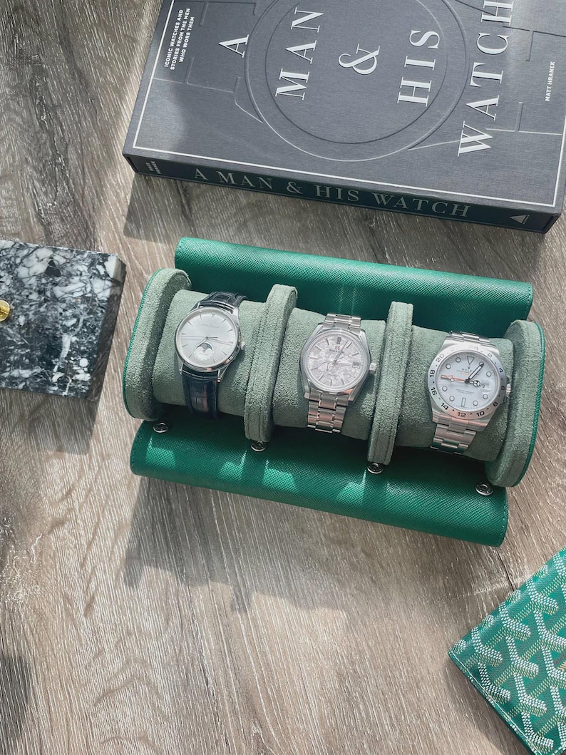 May include: A green leather watch roll holding three luxury watches with silver and black watch faces. A book titled "A Man & His Watch" is visible in the background. A marble case and a green wallet are also in the image.