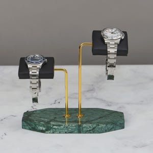 Könnte beinhalten: Ein Uhrenständer mit zwei Armbanduhren. Der Ständer hat einen grünen Marmorsockel, goldfarbene Metallstützen und schwarze Uhrenkissen. Eine Uhr hat ein blaues Zifferblatt, die andere ein weißes Zifferblatt.