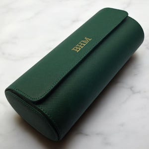 May include: A dark green, cylindrical travel case with a flap closure. The case has gold-colored initials "BHM" on the front. The case is made of a textured material and is resting on a marble surface.