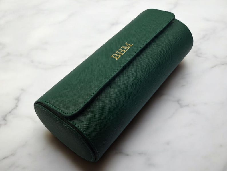 May include: A dark green, cylindrical travel case with a flap closure. The case has gold-colored initials "BHM" on the front. The case is made of a textured material and is resting on a marble surface.