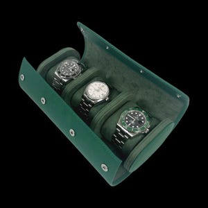 May include: A green leather watch travel case with three compartments, each holding a silver wristwatch. The case is open, revealing the watches against a green felt interior. The case is secured with silver snap closures.