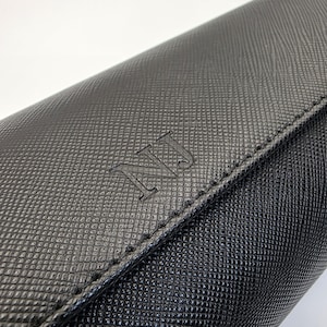 May include: Close-up of a black leather wallet with a textured pattern. The wallet has a flap closure and the initials "NJ" embossed on the front. The stitching is visible along the edges.