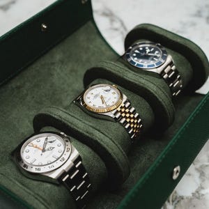 May include: Four luxury wristwatches displayed in a green velvet travel case. The watches have silver and gold bands, white faces, and black or blue bezels. The case is open, revealing the watches nestled in individual compartments.