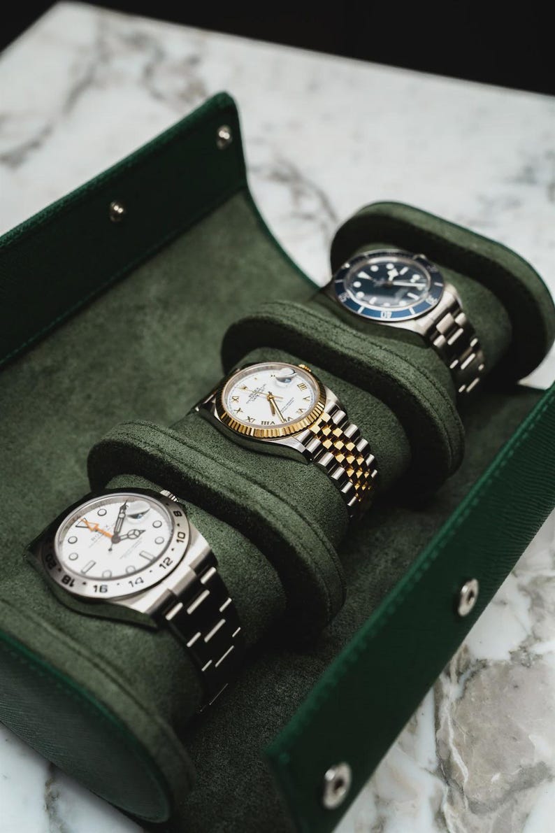 May include: Four luxury wristwatches displayed in a green velvet travel case. The watches have silver and gold bands, white faces, and black or blue bezels. The case is open, revealing the watches nestled in individual compartments.