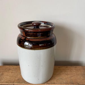 May include: A ceramic jar with a lid. The jar is white with dark brown accents around the top and lid. The jar is sitting on a wooden surface. The jar is a vintage style.