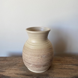 May include: A ceramic vase with a rounded body and a flared rim. The upper portion features a smooth, light cream glaze, while the lower section displays a textured, horizontal ribbed pattern with earthy brown and tan tones throughout the surface.