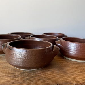 May include: A set of five round brown ceramic bowls, each featuring two small side handles. The bowls have a textured, dark brown glazed exterior with a smooth interior finish and a light-coloured base visible at the bottom of each vessel.