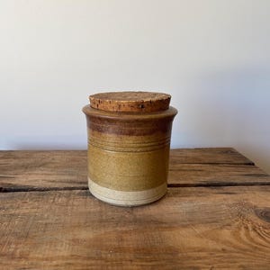 May include: A cylindrical stoneware canister featuring a multi-toned brown and tan glaze with horizontal incised lines around the upper body. The container is topped with a circular, textured natural cork lid that fits securely into the rim.