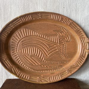 May include: Oval brown ceramic tray with a raised relief design of a stylized animal figure. The tray has two small handles and a textured border. The animal figure is centered on the tray.