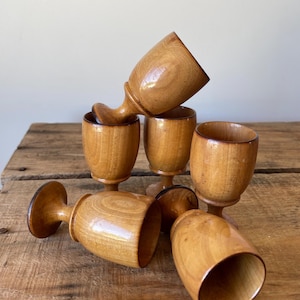 May include: A collection of six small wooden goblets with a smooth, polished finish. Each goblet features a rounded bowl, a short stem, and a circular base. The wood displays natural grain patterns in varying shades of light and medium brown.